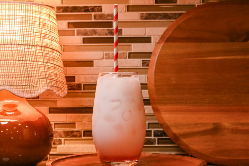 A pink drink in a glass with a striped straw sitting on a kitchen counter with a lamp and cutting board in the background.