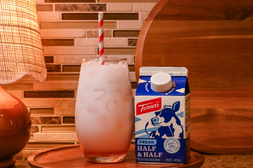 A pink drink in a glass with a striped straw sitting on a kitchen counter next to a pint of Turner's Half & Half. A lamp and cutting board  are in the background.