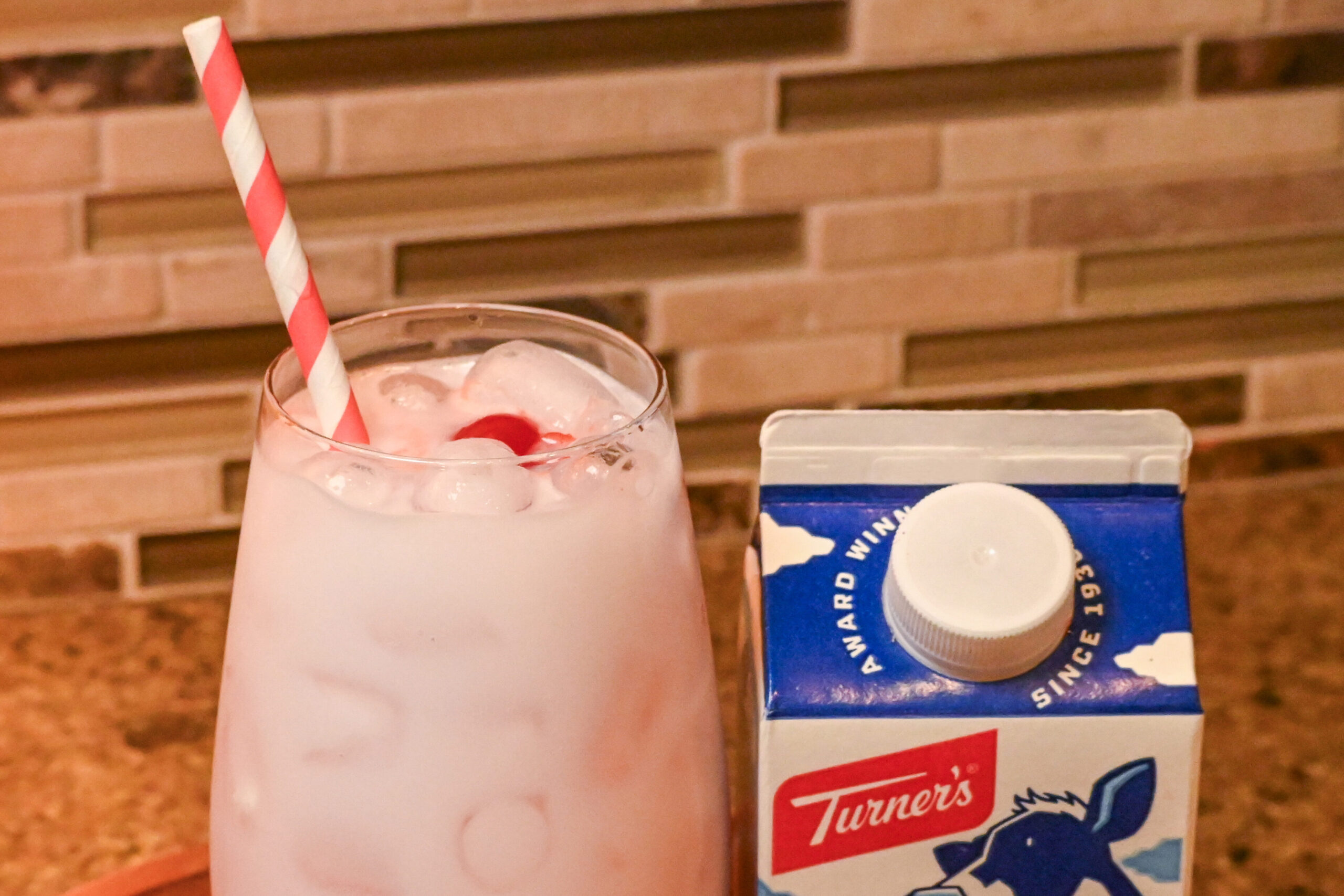 A closeup image  in a kitchen setting of a pink drink in a glass with a striped straw next to a pint of Turner's Half & Half.