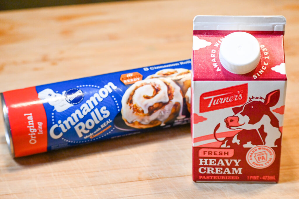 A package of Pillsbury Cinnamon Rolls behind a pint of Turner's Heavy Cream on a wooden cutting board.