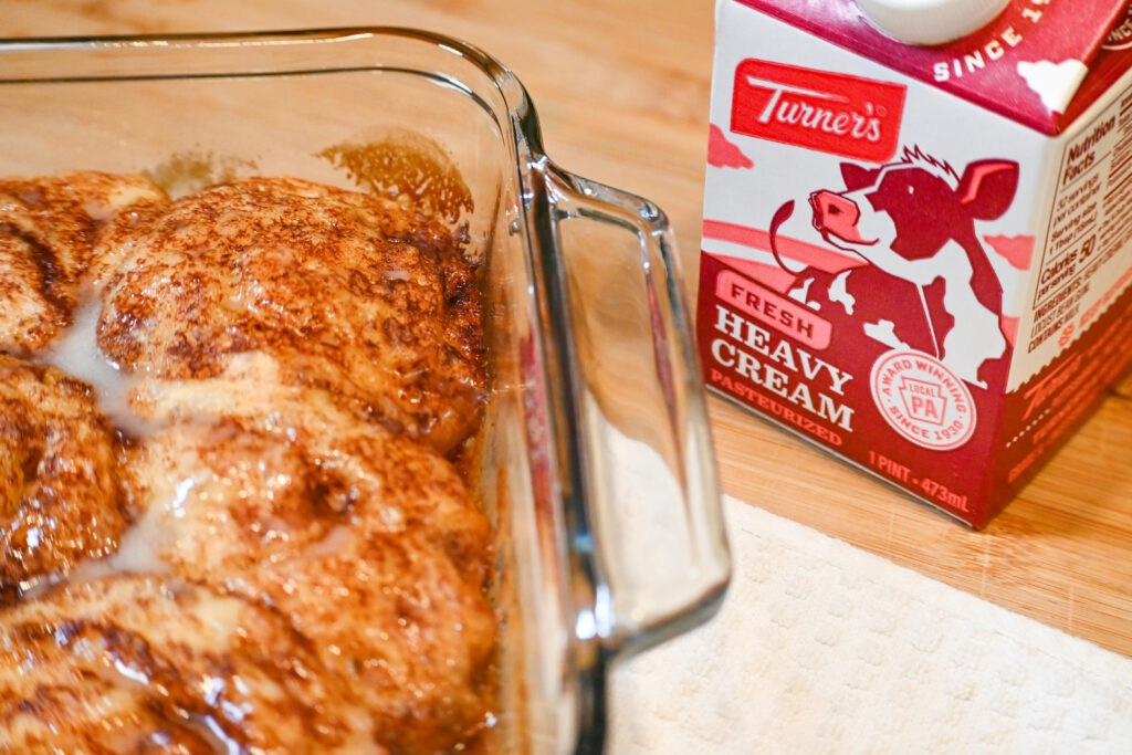 Cinnamon Rolls angled in a glass baking dish with a pint of Turner's Heavy Cream beside it on  a wooden cutting board.