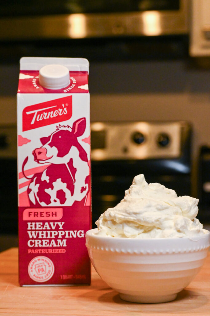 A court of Turner's Heavy Cream and a small bowl of whipped cream sit on a wooden cutting board with an out of focus oven in the background.